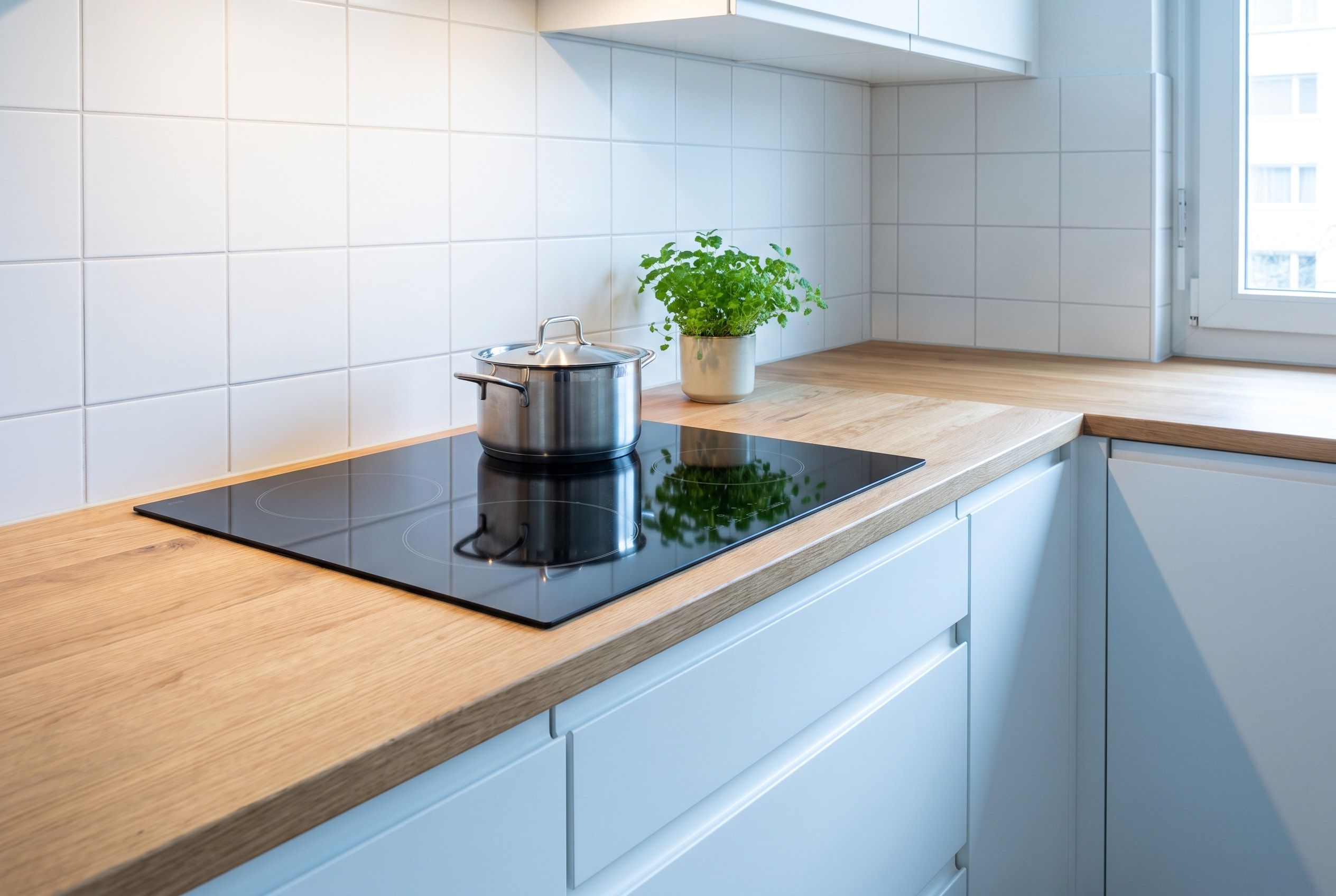 Table de cuisson à induction noire encastrée dans un plan de travail en bois clair, casserole inox et pot d’herbes aromatiques