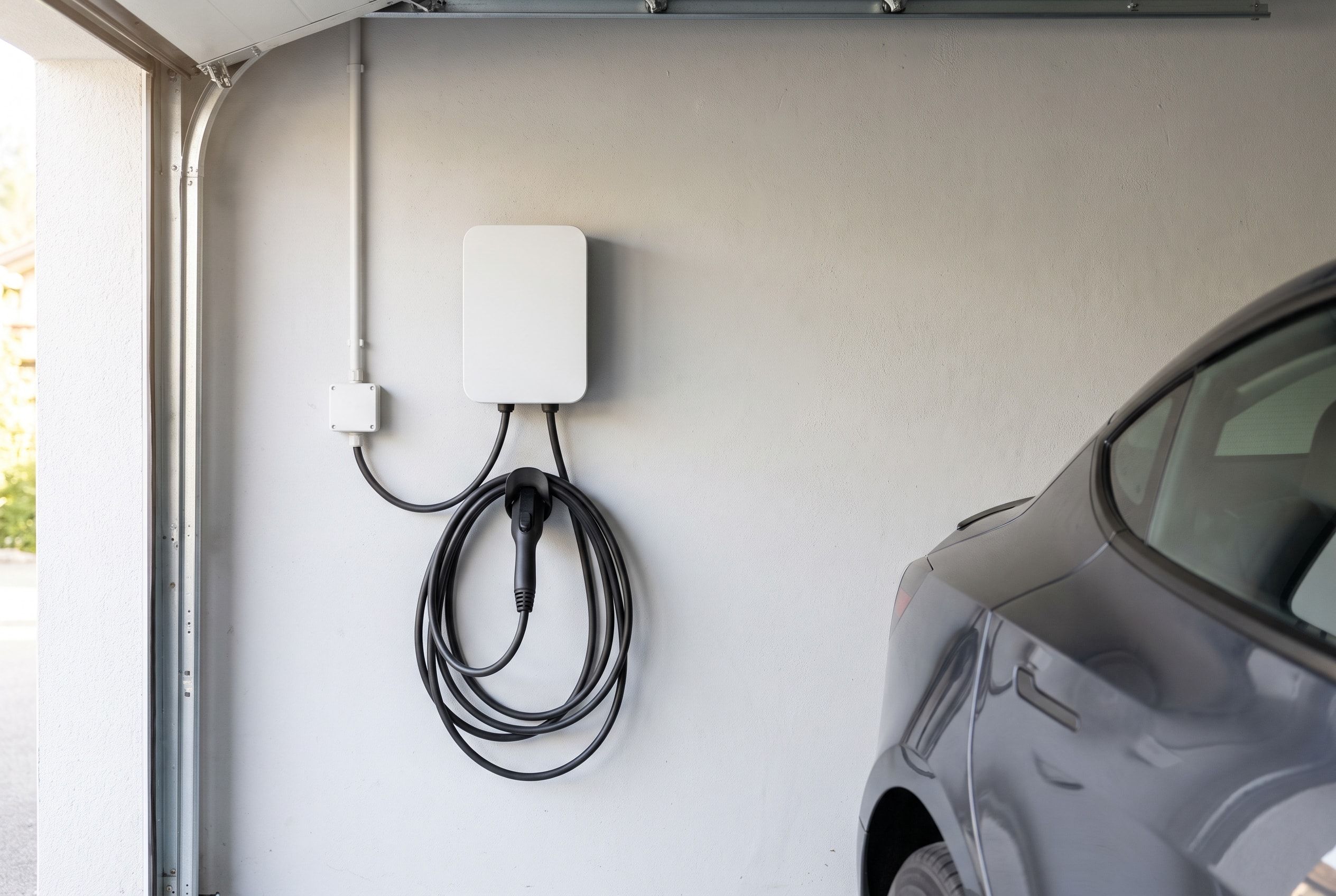 Borne de recharge blanche fixée au mur d’un garage, câble de charge noir enroulé, voiture électrique grise garée à côté